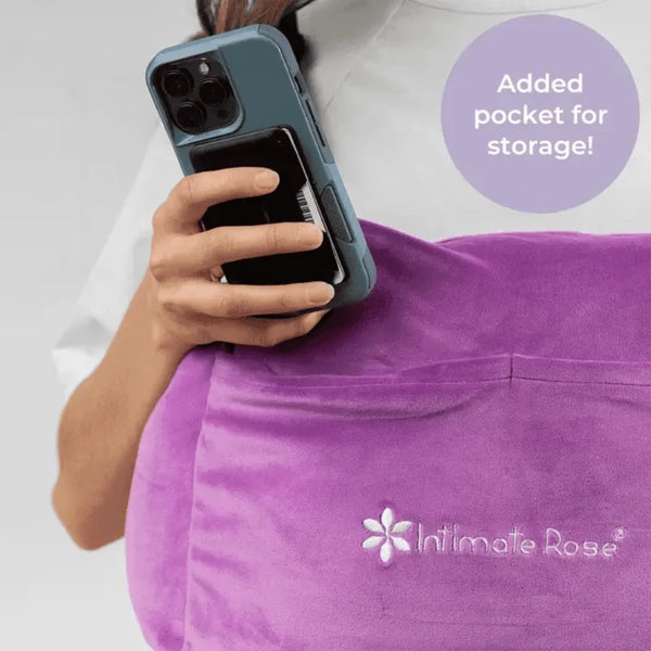 A person is holding a smartphone with a teal case, slipping it into a small pocket on the purple Post-Surgery Mastectomy Pillow by Intimate Rose. A text bubble says, "Added pocket for storage!" The product features a floral logo.