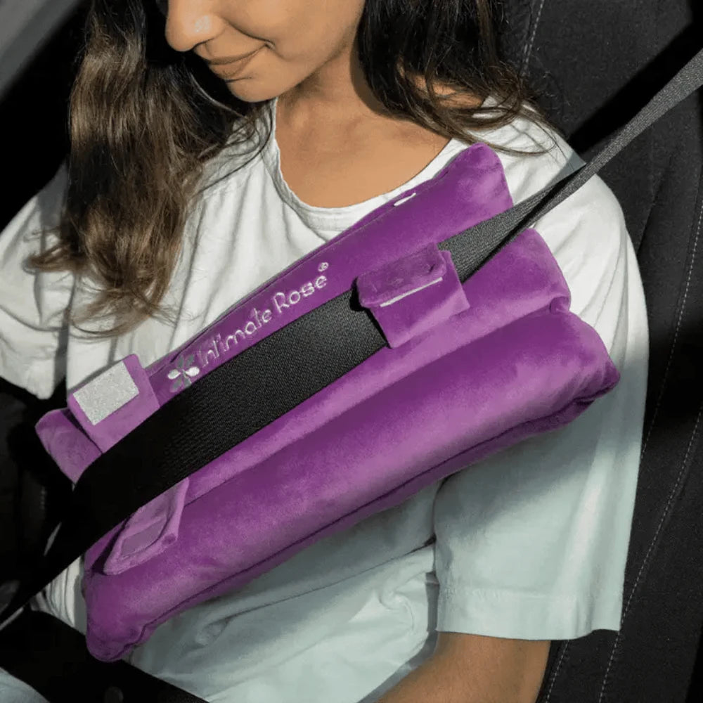 A person seated in a car uses the Intimate Rose Post-Surgery Seat Belt Pillow to enhance comfort after surgery. This cushion, akin to a mastectomy pillow, attaches to the seatbelt and offers pain relief as they rest their arm on it. The individual is wearing a white shirt, with the photograph capturing part of their upper body.