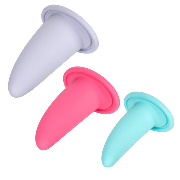 Three silicone therapy dilators in varying sizes and colors are laid out on a white background. The largest is light gray, the medium one is pink, and the smallest is light blue. The She-ology™ 3-piece Wearable Vaginal Dilator Set by Calexotics, made from body-safe silicone, features a flared base for easy handling during vaginal dilation exercises.