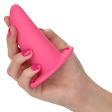 A hand with pink-painted nails holds a flared-base plug from the She-ology™ 3-piece Wearable Vaginal Dilator Set by Calexotics. The fingers grip the cylindrical dilator, which tapers toward the top. Made of body-safe silicone, it stands out against the plain white background.