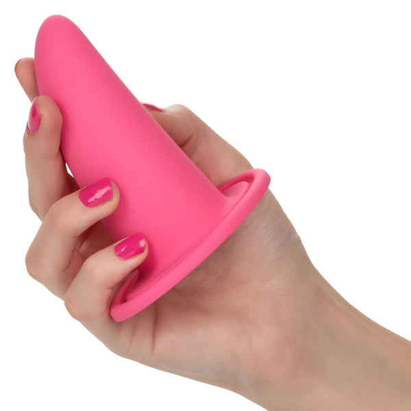 A hand with pink-painted nails holds a flared-base plug from the She-ology™ 3-piece Wearable Vaginal Dilator Set by Calexotics. The fingers grip the cylindrical dilator, which tapers toward the top. Made of body-safe silicone, it stands out against the plain white background.