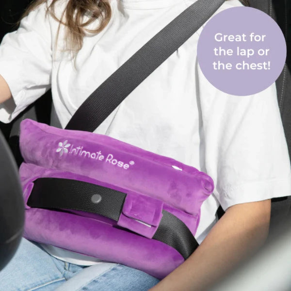 A person sporting a white shirt is using an Intimate Rose Post-Surgery Seat Belt Pillow in purple as a seatbelt cushion in their car. The text on the image states, "Great for post-surgery comfort—perfect for the lap or chest!