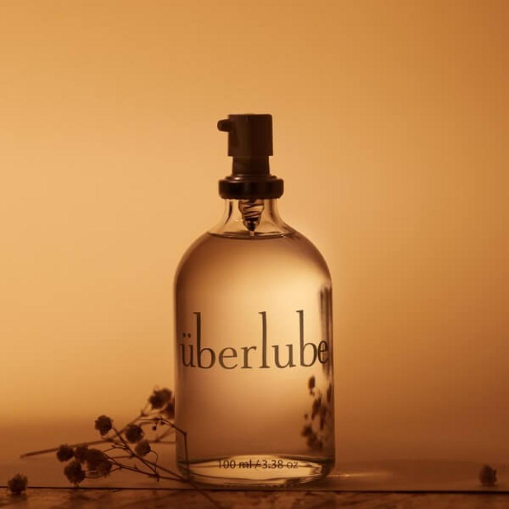 A 100 ml (3.38 oz) glass bottle with a pump dispenser, branded as "Uberlube," is placed centrally against a warm amber-toned backdrop. Accompanying this premium silicone lubricant, dried flowers embellish the scene, highlighting its role in enhancing sexual wellness.