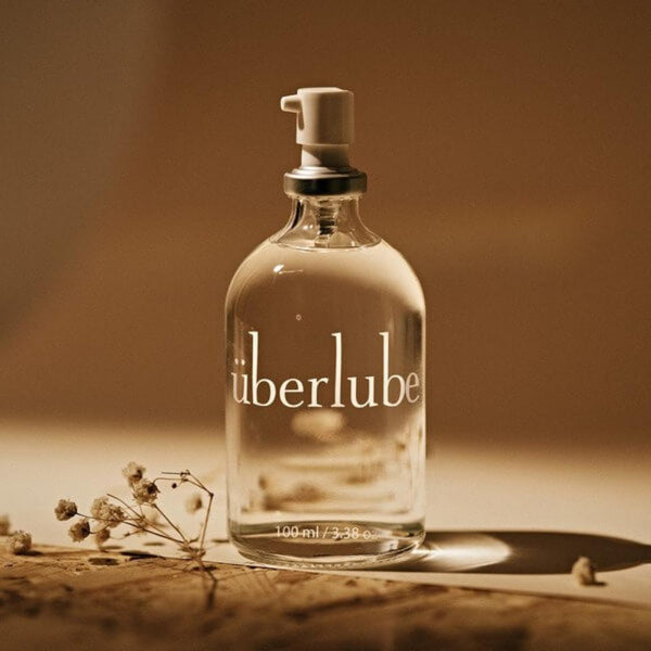 A glass bottle of Uberlube rests on a smooth surface. Featuring a pump dispenser and "100 ml / 3.38 oz" text, it exudes elegance. Soft, neutral lighting and dried flowers enhance the ambiance, creating a scene dedicated to sexual wellness.
