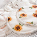 Close-up of three circular, reusable breastfeeding pads adorned with soft, floral designs. One pad showcases sunflowers, while the others display different patterns. The organic bamboo nursing pads rest on a white, textured surface. The brand name 
