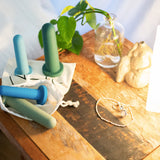 A wooden bedside table features the Bien Silicone Vaginal Dilators - Large Set by BIEN Australia, ideal for vaginal dilator therapy, along with three colorful silicone dumbbells. Additionally, a small green plant in a glass vase, a soft lamp, and silver jewelry are placed on the surface, collectively promoting aspects of women's health.