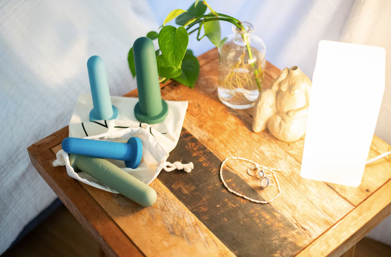 A wooden bedside table features the Bien Silicone Vaginal Dilators - Large Set by BIEN Australia, ideal for vaginal dilator therapy, along with three colorful silicone dumbbells. Additionally, a small green plant in a glass vase, a soft lamp, and silver jewelry are placed on the surface, collectively promoting aspects of women's health.