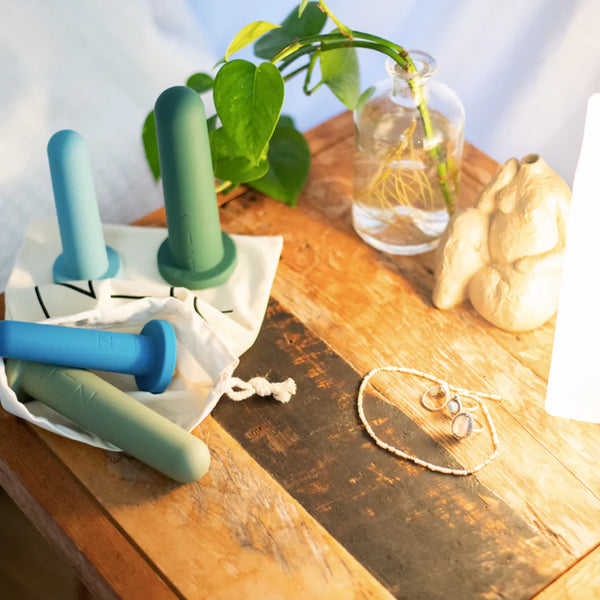 A wooden bedside table features the Bien Silicone Vaginal Dilators - Large Set by BIEN Australia, ideal for vaginal dilator therapy, along with three colorful silicone dumbbells. Additionally, a small green plant in a glass vase, a soft lamp, and silver jewelry are placed on the surface, collectively promoting aspects of women's health.