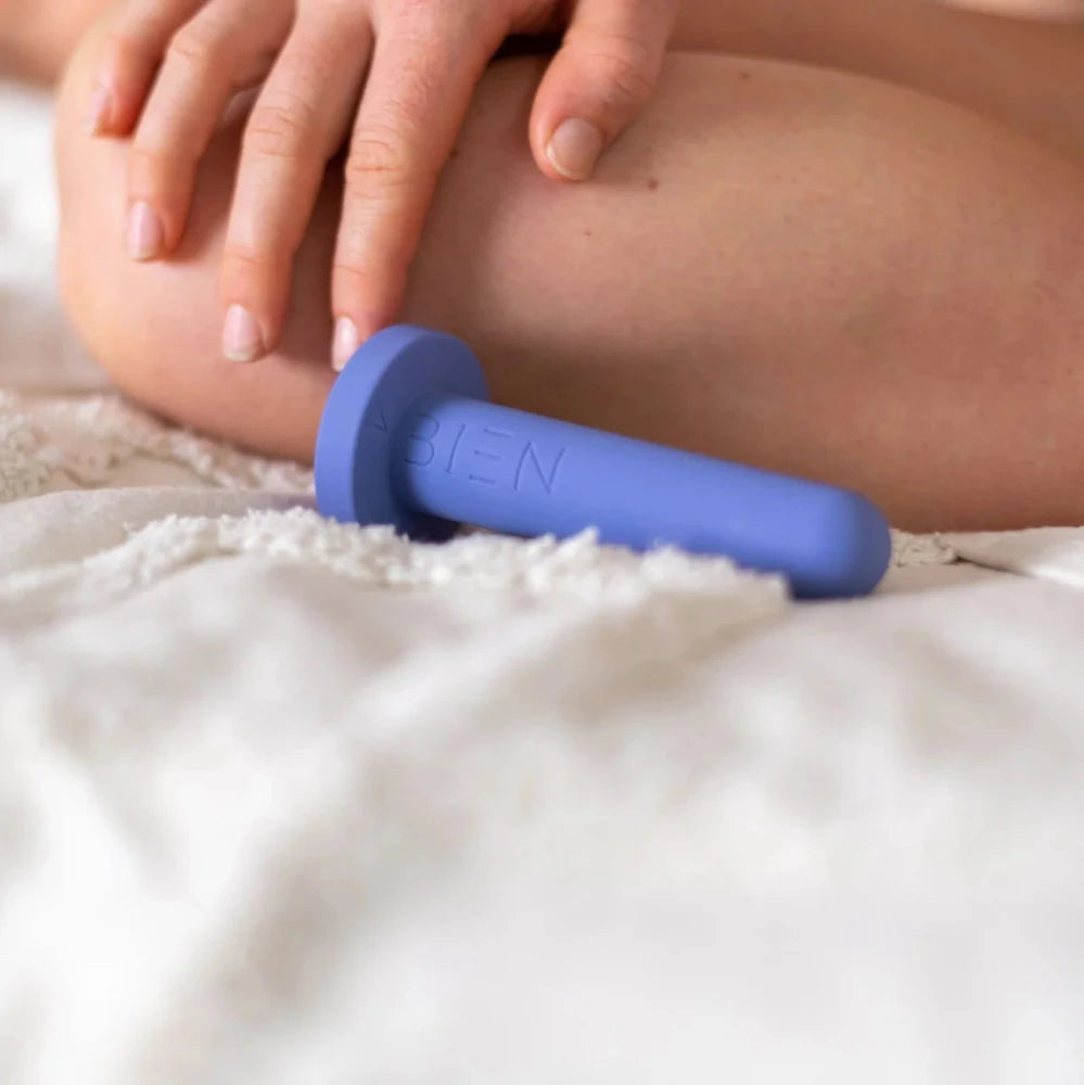 A Bien Silicone Vaginal Dilator from the Small Set by BIEN Australia, crafted from medical-grade silicone in a purple hue, rests on a white textured surface. In the background, a person with light skin gently places their hand on their leg, evoking a sense of calm and comfort.