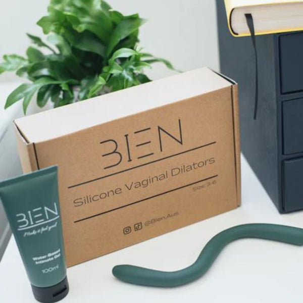 A cardboard box labeled "Silicone Vaginal Dilators" from BIEN Australia sits on a white surface. Next to it, there's a green tube of water-based lubricant and The Bien Wave - Pelvic Floor Wand in green for pelvic floor muscle training. In the background, there's a plant and a black, bound book.