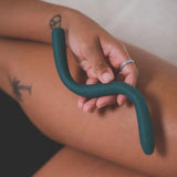 A person wearing a ring on their finger holds The Bien Wave - Pelvic Floor Wand by BIEN Australia, which is dark green and curved, against their thigh that has a tattoo of a palm tree and peace sign. The background appears to be white fabric.