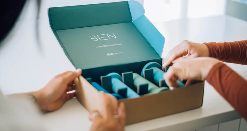 Two individuals are opening a BIEN-branded cardboard box containing several teal-colored, cylindrical items, possibly skincare or beauty products. The box interior is also teal, creating a cohesive color scheme. The scene is set on a white surface.