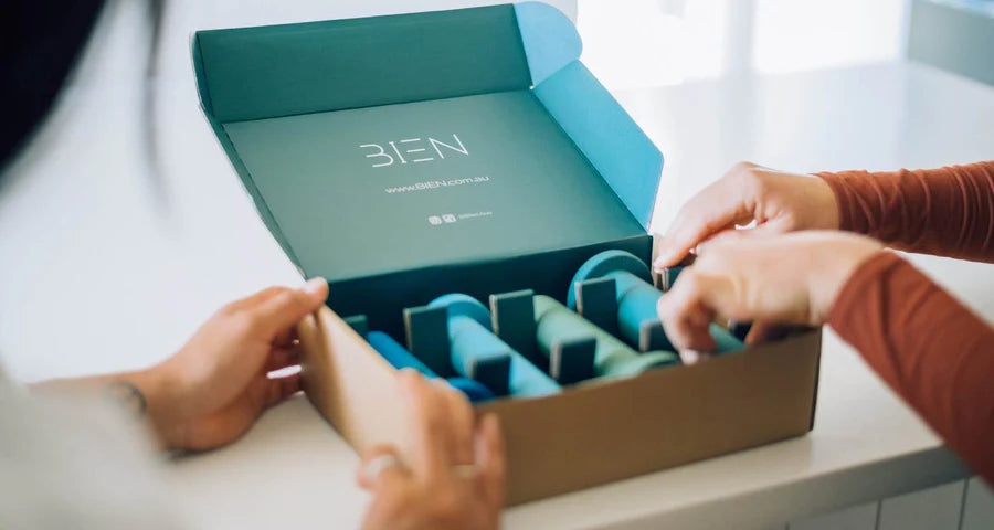 Two individuals are opening a BIEN-branded cardboard box containing several teal-colored, cylindrical items, possibly skincare or beauty products. The box interior is also teal, creating a cohesive color scheme. The scene is set on a white surface.