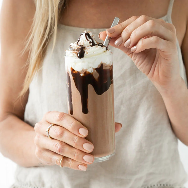 A woman wearing a beige shirt holds a tall glass filled with Lactation Hot Chocolate with Collagen from The Breastfeeding Tea Co, topped with whipped cream and chocolate syrup. She stirs the drink with a metal straw. The focus is on the hot chocolate and her hands, which display neatly manicured nails.