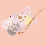 A mesh bag containing Bubba Bump Organic Bamboo Reusable Breast Pads - 14 Pack and a tag displaying product information. The ultra-absorbent pads feature various designs, including floral and leaf patterns. The bag and pads are set against a soft peach background, with dried grasses placed beside them.