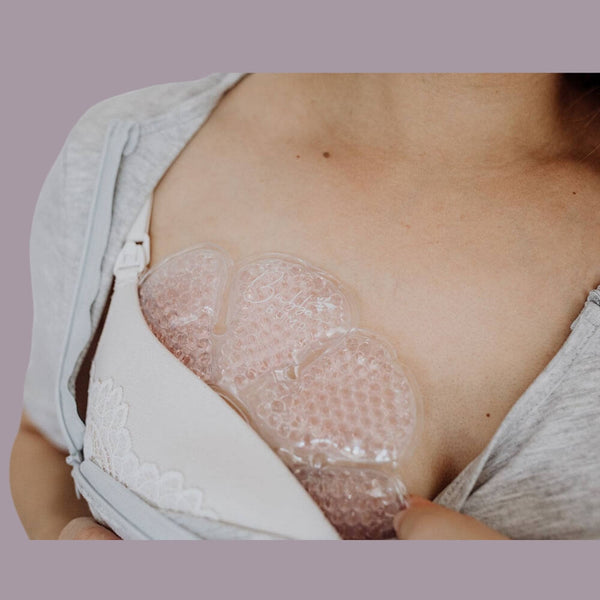 A person is shown applying a Bubba Bump Ice And Heat Pack For Breasts inside the cup of a white and gray lace-detailed bra, which features an open flap to reveal the mastitis relief pad. The background is a solid light mauve color.