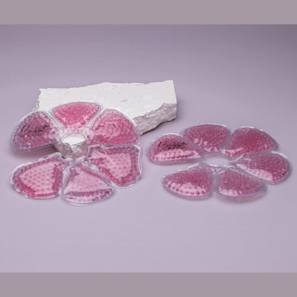 Two Bubba Bump "Ice And Heat Pack For Breasts" gel bead therapy pads in pink, each divided into six sections, lie on a light purple surface. One pad is draped over a textured white foam block for mastitis relief, while the other rests flat beside it as an effective breast ice pack.