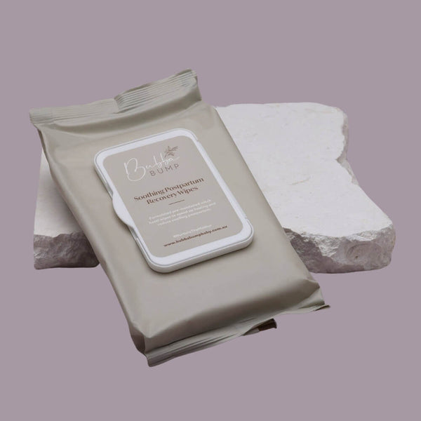 A beige packet of "Bubba Bump Postpartum Witch Hazel Recovery Wipes // 45 Pack" rests on a white stone surface against a light gray background. The packet features a label with the product name and some additional text in a clean, modern font.