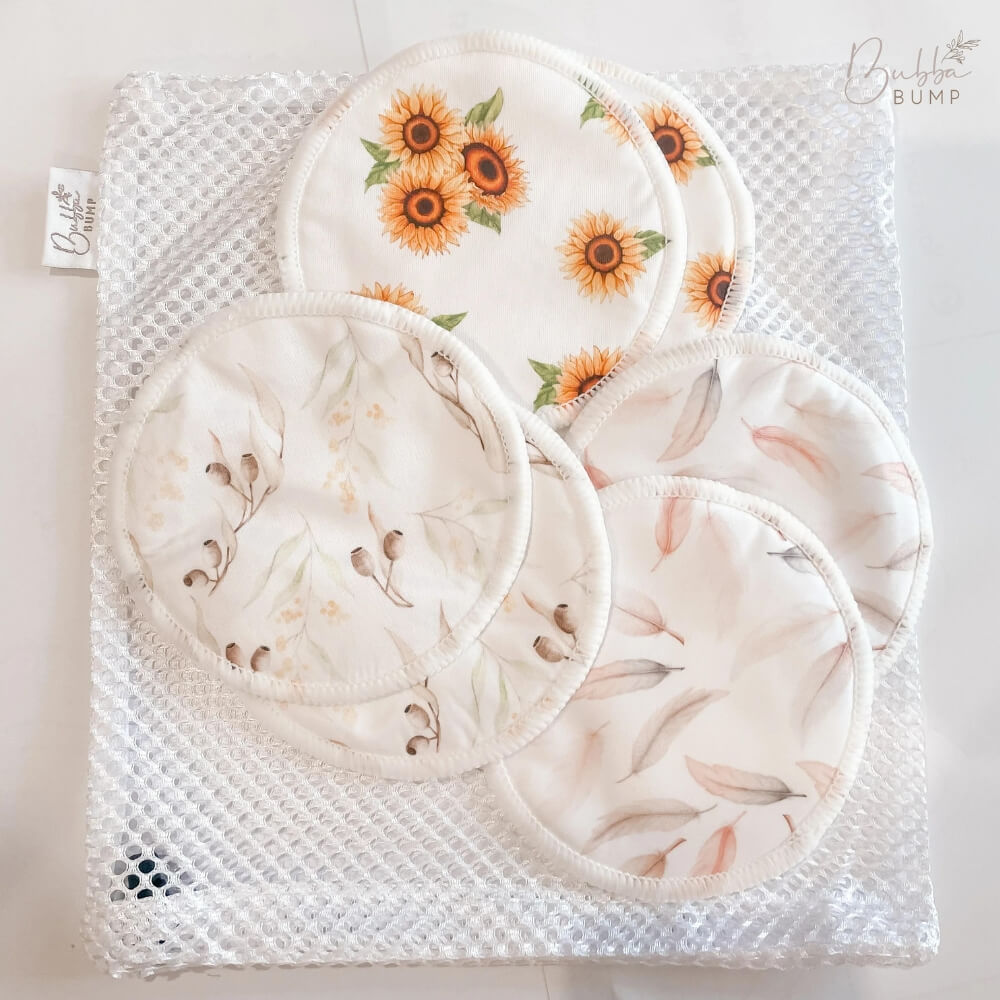 A set of round, ultra-absorbent breast pads with various floral and leaf patterns in soft colors (sunflowers and light foliage designs) are neatly arranged on a white mesh laundry bag. The brand name "Bubba Bump" is visible in the corner of the image.