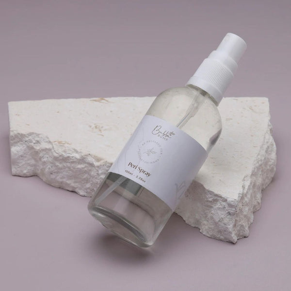 A spray bottle of Bubba Bump's Postpartum Recovery Perineal Spray with a white label lies on a piece of white textured stone against a light mauve background. The translucent bottle features a white cap and spray nozzle. The arrangement is minimalist and aesthetically pleasing.
