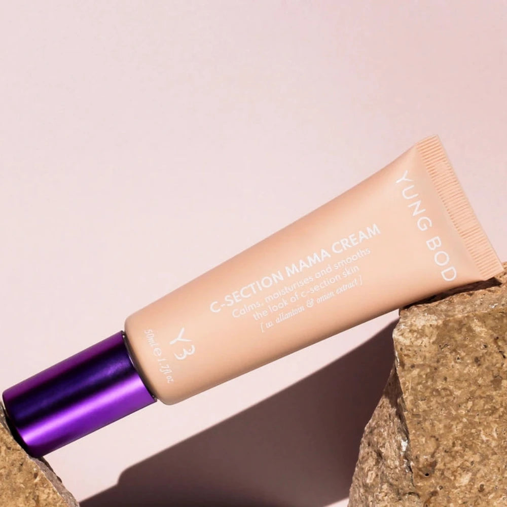 A tube of Yung Bod's C-Section Mama Cream is displayed between two stones. The cream, made with natural ingredients, is designed to calm, moisturize, and smooth the look of C-section skin. The light beige tube with a metallic purple cap features text detailing its scar-softening properties and usage information.