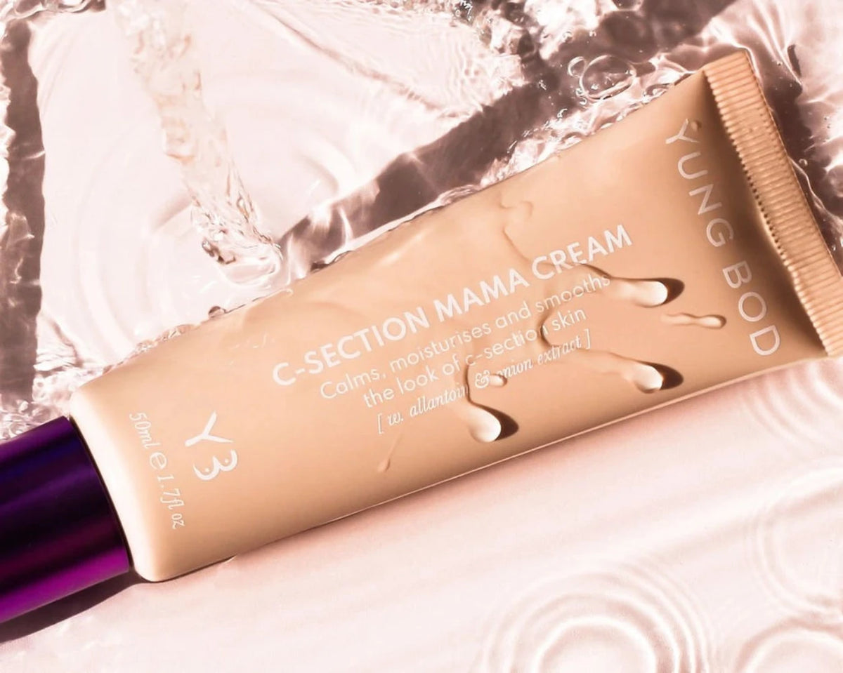 A tube of C-Section Mama Cream from Yung Bod rests on a reflective, watery surface. The beige tube with a purple cap is adorned with water droplets. The text highlights that this C-Section Scar Softening Cream soothes, hydrates, and smoothens the appearance of c-section skin using natural ingredients.