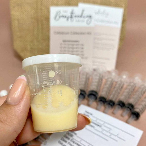 A hand holds a small clear measurement cup containing a creamy yellow liquid. In the background, there are several syringes filled with the same liquid, informational cards, and a burlap pouch. The setting suggests **The Breastfeeding Tea Co's Antenatal Colostrum Collection Kit** designed for colostrum collection during breastfeeding or antenatal periods.