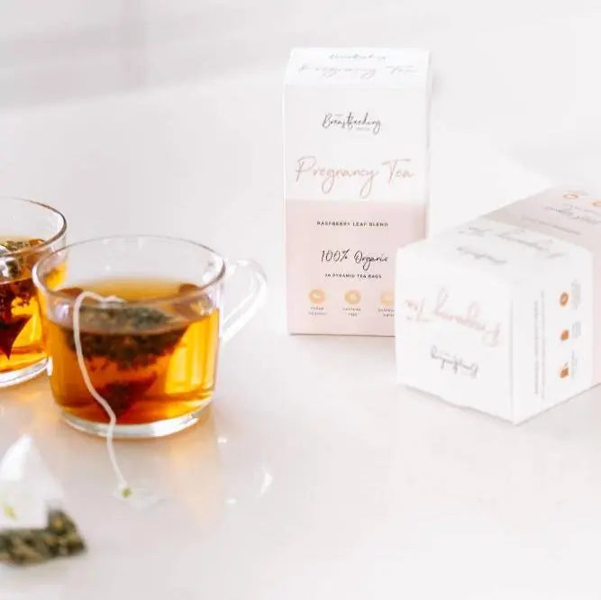 Two glass cups with tea bags sit beside a white "Pregnancy Tea - Raspberry Leaf Tea" box from The Breastfeeding Tea Co, featuring pregnancy-safe herbs. Another tea bag and a second box are also nearby.
