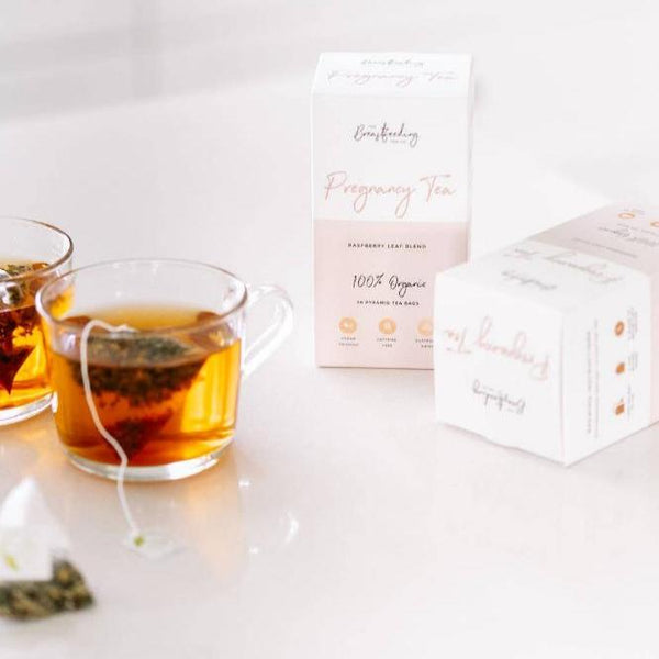 A box of "Pregnancy Tea - Raspberry Leaf Tea" by The Breastfeeding Tea Co, labeled as "Raspberry Leaf Blend, 100% Organic," sits on a reflective white surface, partially opened with some tea bags visible. Beside it are two glass cups filled with Pregnancy Tea; one has a tea bag submerged while the other has a tea bag placed next to it.