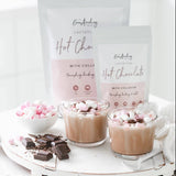Two glass mugs filled with The Breastfeeding Tea Co's Lactation Hot Chocolate with Collagen topped with marshmallows sit on a white table. Next to the mugs are chunks of chocolate and a bowl of mini marshmallows. Behind the mugs, two packets labeled 