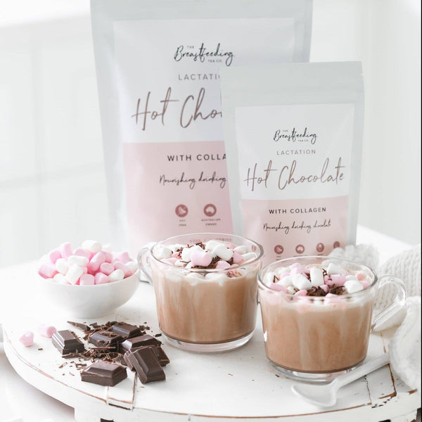 Two glass mugs filled with The Breastfeeding Tea Co's Lactation Hot Chocolate with Collagen topped with marshmallows sit on a white table. Next to the mugs are chunks of chocolate and a bowl of mini marshmallows. Behind the mugs, two packets labeled "Lactation Hot Chocolate with Collagen" highlight that this soothing drink supports breastmilk supply.