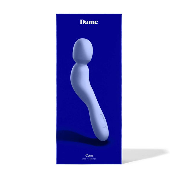 A blue package contains the Dame Com, an ergonomically shaped wand vibrator in gray, featuring a curved design and flexible neck. The packaging prominently showcases the Dame brand name against a dark blue background, promising those signature rumbly vibrations for enhanced pleasure.