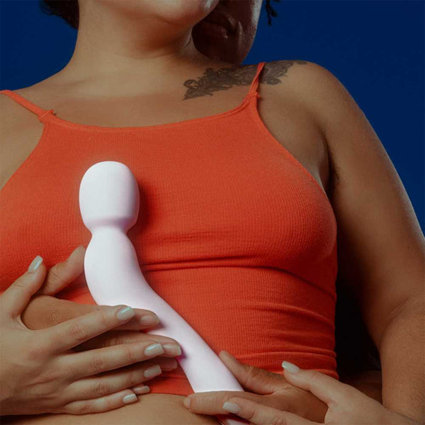 Two individuals are partially visible, with one wearing an orange tank top. They hold the Dame Com - External Wand Vibrator by Dame, featuring a flexible neck, against a person's chest. One person has a visible tattoo on their shoulder, set against a solid blue background.