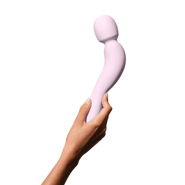 A hand holds the Dame Com - External Wand Vibrator by Dame, a light pink device with rumbly vibrations against a white background. This massager boasts a smooth surface and a flexible neck, highlighting its unique bend for precise use.