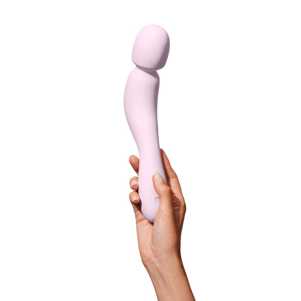 A hand holds a Dame Com - External Wand Vibrator by Dame, showcasing its rumbly vibrations and light pink, flexible neck elegantly curved against a white background.