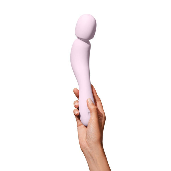 A hand holds a Dame Com - External Wand Vibrator by Dame, showcasing its rumbly vibrations and light pink, flexible neck elegantly curved against a white background.