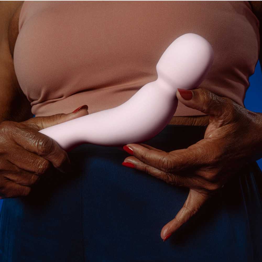 An individual holds the Dame Com - External Wand Vibrator by Dame in a pink shade, its flexible neck enhancing comfort, against a neutral backdrop. They are dressed in a sleeveless top, with their red nail polish adding a vibrant touch.
