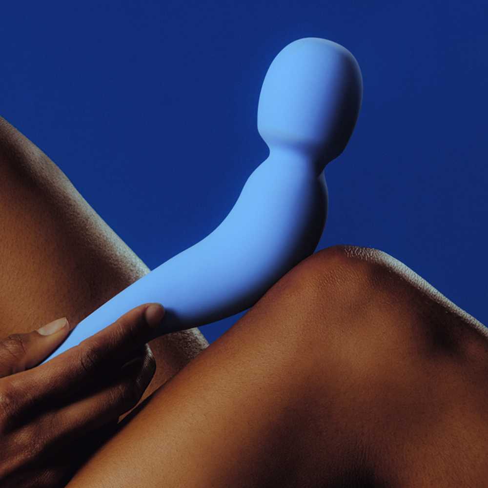 A person holds the Dame Com - External Wand Vibrator by Dame against their knee. The background is a solid blue, which complements both the vibrator's color and its flexible neck. This scene highlights the contrast between skin tone and shades of blue, enhancing the experience of its rumbly vibrations.