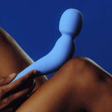 A person holds the Dame Com - External Wand Vibrator by Dame against their knee. The background is a solid blue, which complements both the vibrator's color and its flexible neck. This scene highlights the contrast between skin tone and shades of blue, enhancing the experience of its rumbly vibrations.
