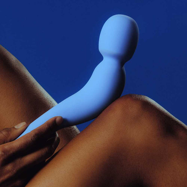 A person holds the Dame Com - External Wand Vibrator by Dame against their knee. The background is a solid blue, which complements both the vibrator's color and its flexible neck. This scene highlights the contrast between skin tone and shades of blue, enhancing the experience of its rumbly vibrations.