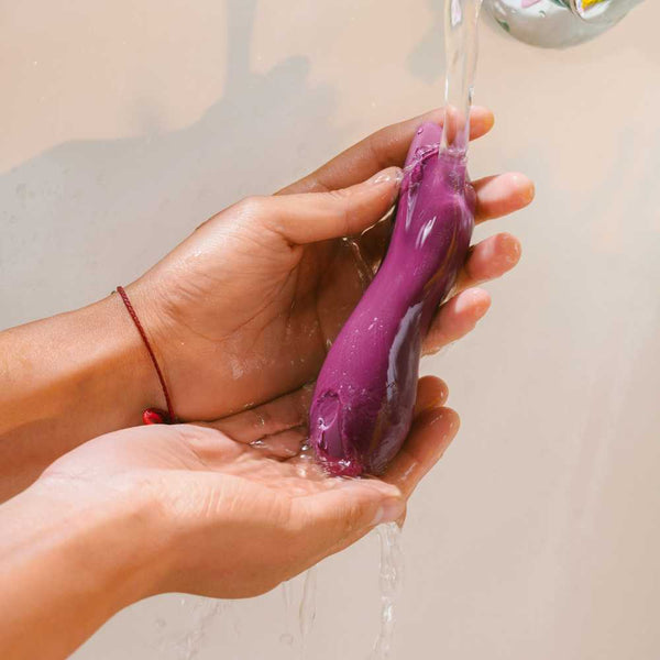 Hands holding the Dame Dip 2.0 - Basic Vibrator, a purple, curved object made of medical-grade silicone from Dame, under running water from a faucet, with water streaming over it.