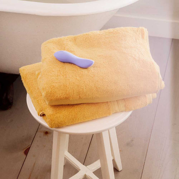 A stack of two yellow towels is neatly placed on a white stool, topped with the Dame Dip 2.0 - Basic Vibrator, crafted from medical-grade silicone. In the background, a white bathtub is partially visible, creating a serene scene set on a wooden floor.
