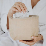 A person dressed in a white robe is holding a beige woven toiletry bag with a zipper. The bag is open, and the individual is removing a transparent plastic-wrapped item from an Antenatal Colostrum Collection Kit by The Breastfeeding Tea Co. They have light pink nail polish and are wearing a silver ring.