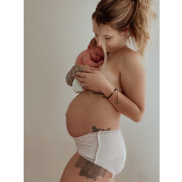 A tattooed woman tenderly cradles a newborn against her chest, wearing Bubba Bump’s Disposable Postpartum Underwear. She stands against a softly lit, plain background, exuding affection and trust in the leak-proof protection provided by Bubba Bump.