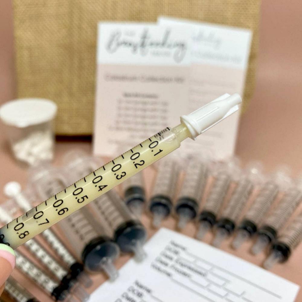 A close-up of a hand holding a small syringe with a needle cap. In the background, several identical syringes and a pile of documents with visible text are on a table. A cylindrical container with small white objects, part of The Breastfeeding Tea Co's Antenatal Colostrum Collection Kit for antenatal expression and breastfeeding, and a burlap-like item are also visible.