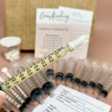 A close-up of a hand holding a small syringe with a needle cap. In the background, several identical syringes and a pile of documents with visible text are on a table. A cylindrical container with small white objects, part of The Breastfeeding Tea Co's Antenatal Colostrum Collection Kit for antenatal expression and breastfeeding, and a burlap-like item are also visible.