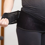 A person in dark leggings and a top fastens the GRDL Pregnancy Belt by GRDL around their lower torso, adjusting the Velcro strap for trusted pelvic and back support during pregnancy.