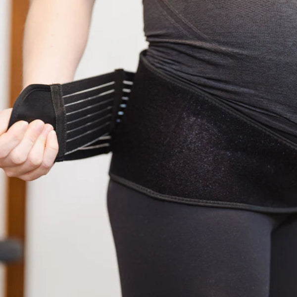 A person in dark leggings and a top fastens the GRDL Pregnancy Belt by GRDL around their lower torso, adjusting the Velcro strap for trusted pelvic and back support during pregnancy.