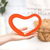 A hand holds the Intimate Rose Bendable Pelvic Wand, a heart-shaped red device designed for pelvic pain relief. The background highlights a serene setting with a white tiled wall, a white vase containing greenery, and a woven basket—perfect for those seeking pelvic floor therapy.