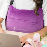 An individual is working at a laptop while wearing a vibrant purple Intimate Rose Post-Surgery Mastectomy Pillow. A blooming pink lily sits in the foreground on the table.